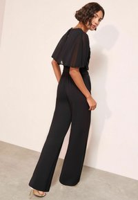 Černý overal s průhlednými, splývavými rukávy a uvolněným střihem. Hladká textura s páskem v pase a širokými kalhotami. Jednoduchý design vzadu.