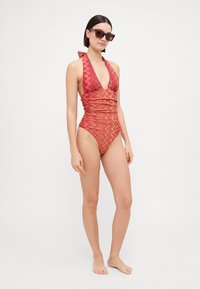 Sarkans un oranžs rakstains viengabala peldkostīms ar haltera kakla dizainu un sakrustotu apdari. Modelis valkā tumšas saulesbrilles. Vienkārša balta fona.