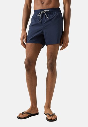 Mandlig model iført marineblå badeshorts med hvid snøre og sorte klipklappere, stående mod en ensfarvet lys baggrund.