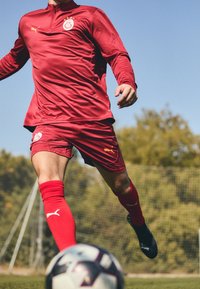 Rød fodbolddragt med lange ærmer og shorts, med et logo på brystet og gyldne detaljer. Spiller i bevægelse, bolden ved foden, græsmark.