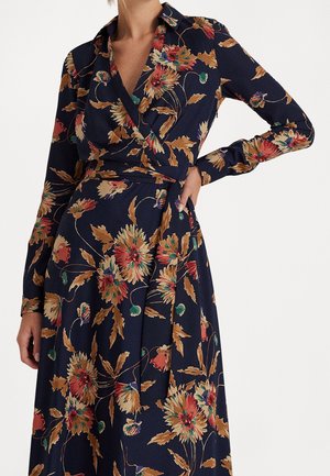 Femme portant une robe portefeuille bleu marine à manches longues avec un motif floral dans des tons orange, rouge, vert et beige.