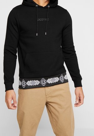 Mężczyzna w czarnej bluzie z kapturem z napisem "UNDEFINED" i geometrycznym szarym wzorem na dole, w połączeniu z beżowymi spodniami, na jednolitym tle.