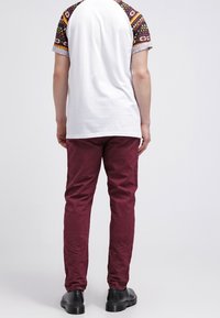 T-shirt blanc avec des manches courtes à motifs, pantalon bordeaux et chaussures noires, vus de dos. Manches texturées avec des motifs géométriques.