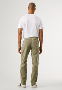 Homem de costas, vestindo uma camisa branca de manga curta, calças verde-oliva e ténis brancos com detalhes verdes.