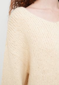 Ελαφρώς μπεζ πλεκτό πουλόβερ με άνετη εφαρμογή, ευρύ λαιμόκοψη, υφασματικό μοτίβο και μαλακό, αφράτο υλικό.