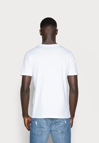 Weißes Baumwoll-T-Shirt mit Rundhalsausschnitt und kurzen Ärmeln, das ein schlichtes Design aufweist. Das Modell trägt blaue Denim-Jeans.