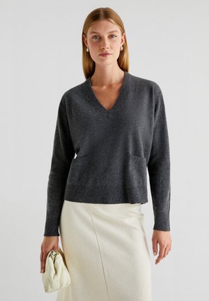 Maglione grigio scuro a V con maniche lunghe, dotato di due tasche frontali e orli a coste, abbinato a una gonna color crema e a una borsa chiara.