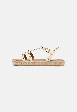 Beige flad sandal med flettet jutesål, tre remme med nitter over foden og en justerbar ankelrem med spænde.