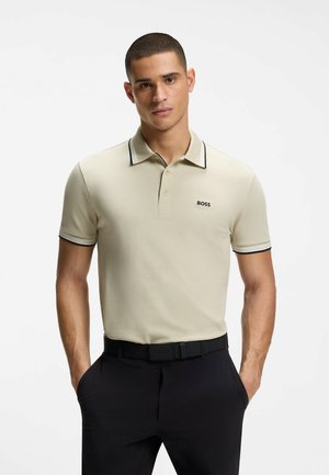 Bărbat cu păr scurt, purtând un tricou polo bej cu logo-ul "BOSS" și pantaloni negri, stând cu mâinile în buzunare pe un fundal simplu.