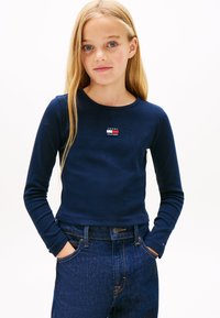 Temno modra majica z dolgimi rokavi z rebrasto teksturo in okroglim izrezom; na prsih majhna večbarvna majica Tommy Hilfiger; kombinirana s temnimi kavbojkami.