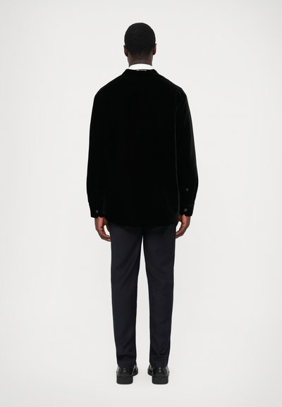 Black velvet overshirt with a collared neck, long sleeves, and buttoned cuffs, paired with navy trousers and black shoes.