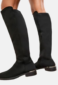 Botas negras de ante hasta la rodilla, con cremallera lateral y un pequeño adorno dorado en el tacón. Textura suave en toda la superficie.