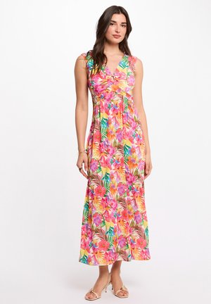 Donna che indossa un vestito senza maniche, con scollo a V, lungo fino alla caviglia, con motivi floreali e foglie multicolori vivaci, abbinato a sandali con tacco beige.