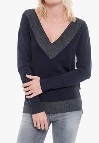 Femme portant un pull noir à col en V avec bordure métallique et un jean gris clair, debout les bras croisés devant.