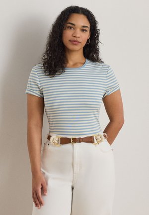 Kvinne med krøllete hår iført blå og hvitstripet kortermet skjorte, hvite bukser og et brunt belte med tau-detaljer, stående mot en ensfarget bakgrunn.