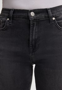 Jean noir taille haute avec une texture lisse, cinq poches et des accessoires en argent, dotés d'une fermeture à bouton et de passants de ceinture.