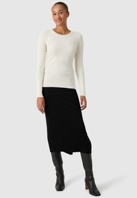 Rippene Langarm-Oberteil in Off-White, kombiniert mit einem schwarzen Strickrock. Schwarze kniehohe Stiefel betonen den Look. Glatte Texturen und figurbetonte Passform.