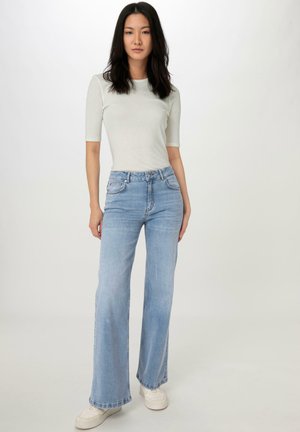 Los jeans acampanados de color azul claro presentan una cintura alta, un leve desgaste y un diseño clásico de cinco bolsillos, combinados con una blusa ajustada de color gris claro.