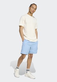 Cream short-sleeve t-shirt with a front logo, paired with light blue shorts and white sneakers with black striped socks. Casual fit.