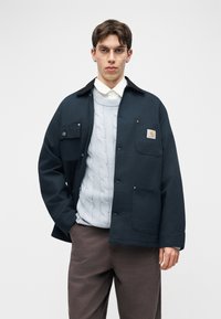 Νέος άνδρας που φοράει διάφανα γυαλιά, ένα σκούρο μπλε σακάκι πάνω από ένα ανοιχτό γκρι πουλόβερ με πλέξη καλώδιου και λευκό πουκάμισο με γιακά, με καφέ παντελόνια μπροστά σε λευκό φόντο.