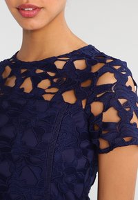 Navy blue dress with floral lace overlay featuring short sleeves and tailored fit. Textured design and open cut-outs on shoulders.