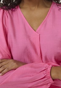 Blouse rose texturée avec un col en V et des manches froncées. Présente une coupe décontractée et des coutures discrètes le long du col.