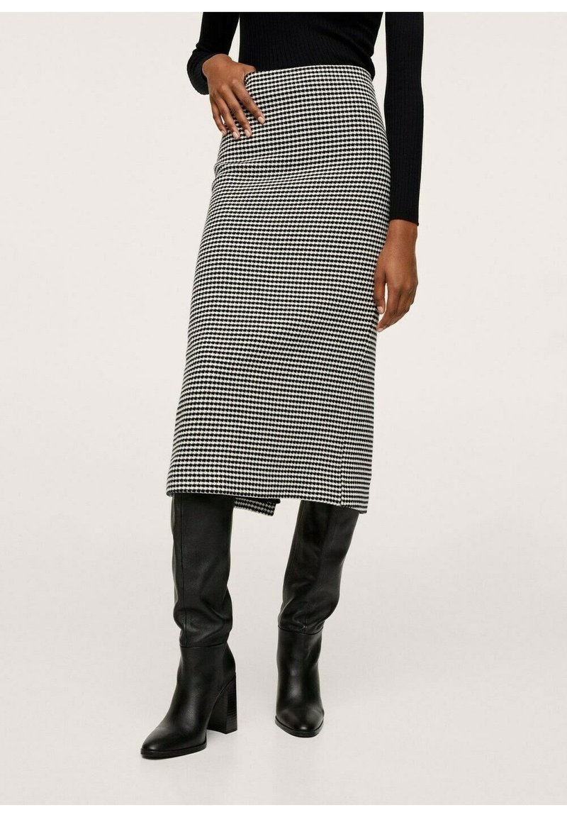 Femme portant une jupe midi pied-de-poule noire et blanche associée à des bottes noires à talons montant jusqu'aux genoux et un haut noir à manches longues.