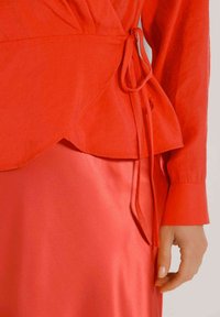 Rotes Wickeloberteil mit einer Knoten-Detail an der Seite, kombiniert mit einem glatten, glänzenden roten Rock. Der Stoff ist weich mit einer matten Oberfläche.