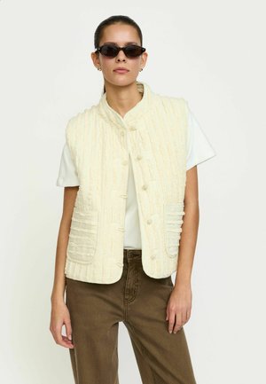 Gilet trapuntato crema con righe verticali, bottoni tondi e due tasche applicate, indossato sopra una maglia bianca e pantaloni marroni.