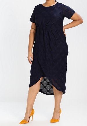 Femme portant une robe midi noire texturée avec des manches courtes et un ourlet asymétrique, associée à des chaussures à talons hauts orange.