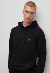 Giovane uomo con capelli corti che indossa un hoodie nero di BOSS con tasca frontale e cordini, in piedi contro un muro grigio semplice.