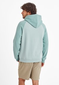Man with curly hair wearing a light blue hooded sweatshirt and beige shorts, facing away against a white background.