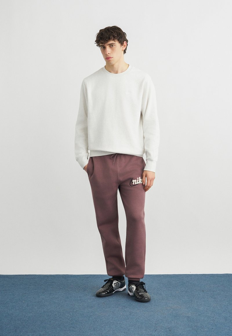 Giovane uomo in piedi che indossa una felpa bianca, pantaloni sportivi Nike color malva con logo e scarpe da ginnastica nere su pavimento blu contro una parete grigia semplice.