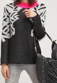 Henkilö, joka pukeutuu musta-valkoiseen kuvioituun neuleeseen, jossa on kirkkaan vaaleanpunainen kaulus, pitää käsissään mustaa teksturoitua käsilaukkua, jossa on kullatut niitit ja hihnat.