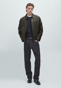 Läderjacka i mörkgrön, marinblå tröja, mörka denimjeans och svarta skor. Rakt skuren silhuett, minimal detaljering, slät textur.