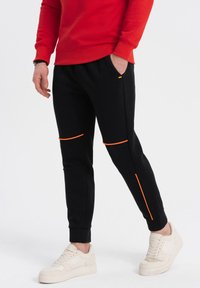 Sorte træningsbukser med orange detaljer, tætsiddende pasform, strukturerede paneler og sidelommer. Parret med lyse sneakers.