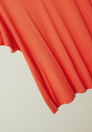 Pomarańczowa koszula z krótkim rękawem wisząca, ukazująca fakturę tkaniny i detale wykończenia na jednolitym jasnym tle.