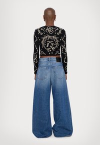 Personne au crâne rasé, vue de dos, portant un haut noir à manches longues avec des motifs de serpent beige et un jean en denim bleu à jambes larges.