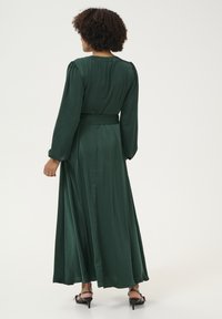 Lungo vestito verde con una texture liscia, caratterizzato da maniche lunghe, una vita aderente con una cintura e una gonna fluida che arriva fino alle caviglie.