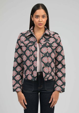 Femme portant une veste noire à motifs floraux rouges et blancs, associée à un jean taille haute foncé et un haut clair en dessous.