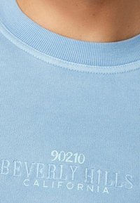 T-shirt di cotone azzurro chiaro con scollo a costine e testo in rilievo "90210 BEVERLY HILLS CALIFORNIA" al centro.