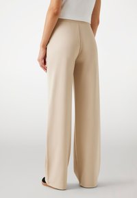 Pantalon large beige en tissu lisse, avec une taille haute, un design plissé et des fentes latérales près de l'ourlet pour un détail supplémentaire.