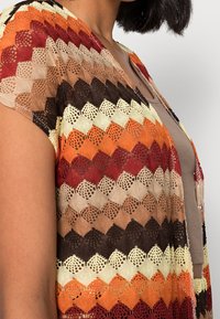 Crocheted open cardigan featuring a zigzag pattern in shades of orange, red, brown, and cream. Lightweight, with short sleeves and a V-neck design.