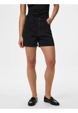 Femeie purtând pantaloni scurți negri din denim cu nituri, top negru și loaferi negri, stând cu o mână în buzunar pe fundal alb.