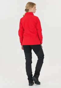 Röd softshell-jacka med hög krage, ihopparad med svarta byxor. Har en figurnära design och bensiddragkedjor, tillverkad av hållbara och väderbeständiga material.