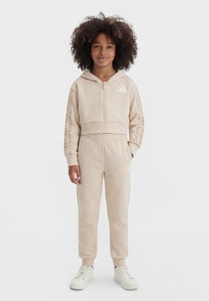 Bambino con capelli ricci che indossa una felpa con cerniera beige e pantaloni da jogging coordinati con sneakers bianche, in piedi contro uno sfondo bianco neutro.