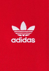Röd tyg med en broderad vit Adidas-logotyp som har tre bladliknande ränder ovanför varumärket i gemener.