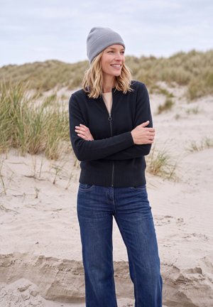 Mujer con gorro gris, chaqueta negra con cremallera y vaqueros azules, de pie con los brazos cruzados sonriendo en una playa de arena con hierba y dunas.