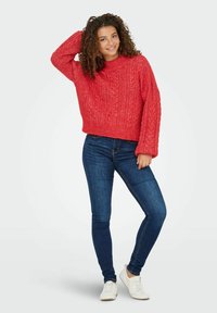 Rood kabelgebreid sweatshirt met een ronde halslijn en lange mouwen, gecombineerd met blauwe skinny jeans en witte sneakers.
