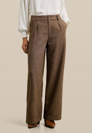 Persoon draagt een bruine wollen broek met hoge taille en wijde pijpen met plooien aan de voorkant, gecombineerd met een witte blouse met lange mouwen en bruine schoenen met hak.